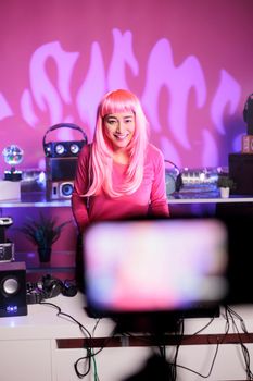 Dj with pink hair performing eletronic song at professional turntables while recording music session with camera, having fun in club during night. Artist dancing while performing techno remix
