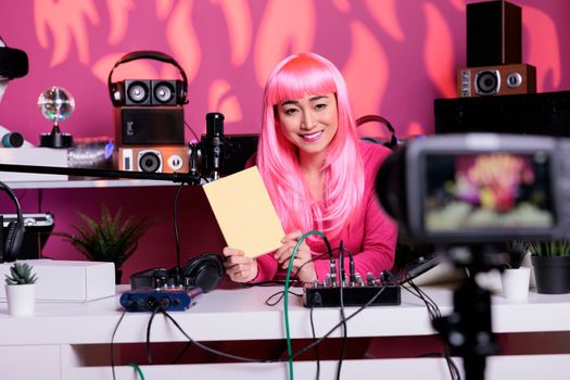 Smiling asian vlogger holding notebook presenting school supplies in front of camera recording review in vlogging studio. Influencer with pink hair filming diary recommendation, live streaming