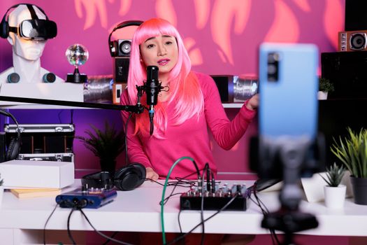Influencer with pink hair talking with subscribers using professional vlogging microphone while recording podcast. Content creator filming vlog with smartphone camera posting on her channel