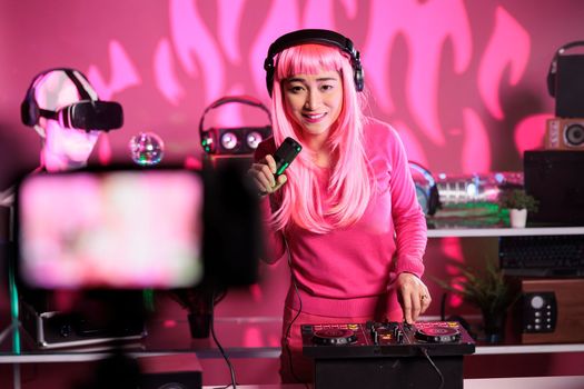 Asian artist playing techno song at mixer console while recording music session using professional camera, preparing to perform in night club. Dj with pink hair using audio equipment