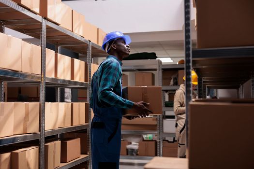 African american storehouse loader holding packed cardboard box and looking at shelf full of containers ready to dispatch. Warehouse employee managing parcels transportation