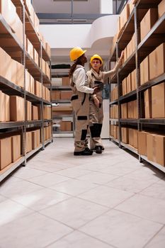 Freight distribution center employees scanning boxes before transportation. Two woman warehouse employees wearing protective helmets and overalls talking, standing near shelf full of cartons