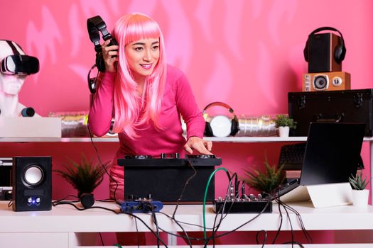 Asian musician standing at dj table enjoying to perform electronic music in front of crowd, using professional turntables in club at night. Performer with pink hair playing techno song