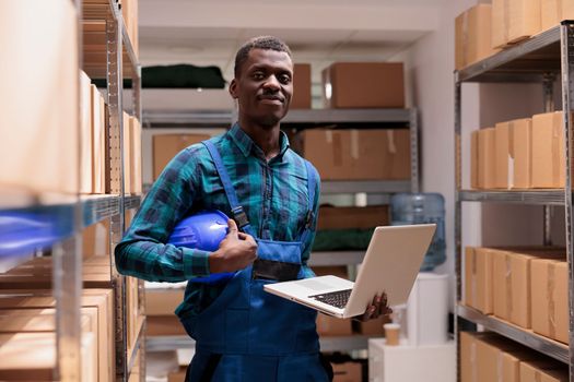 Warehouse employee doing inventory management using laptop, checking parcels before transportation and looking at camera. Smiling african american storehouse worker portrait