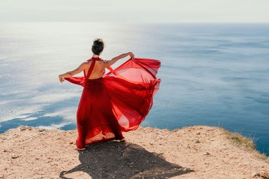 Side view a Young beautiful sensual woman in a red long dress posing on a rock high above the sea during sunrise. Girl on the nature on blue sky background. Fashion photo.