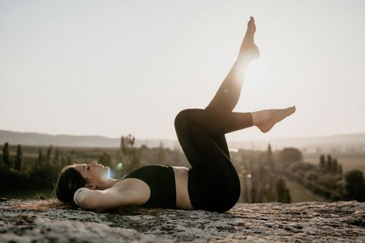Well looking middle aged woman with long hair, fitness instructor in leggings and tops doing stretching and pilates on the rock near forest. Female fitness yoga routine concept. Healthy lifestyle.