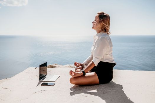 Happy girl doing yoga with laptop working at the beach. beautiful and calm business woman sitting with a laptop in a summer cafe in the lotus position meditating and relaxing. freelance girl remote work beach paradise