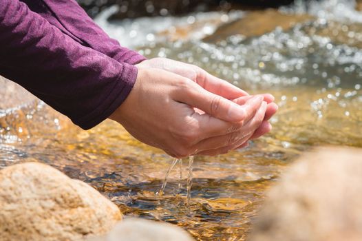 Close-up of water drops falling from female hands into a stream. The hand touches fresh water. A tourist drinks water from a reservoir in the mountains in summer on a sunny day.