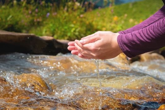 Close-up of water drops falling from female hands into a stream. The hand touches fresh water. A tourist drinks water from a reservoir in the mountains in summer on a sunny day.
