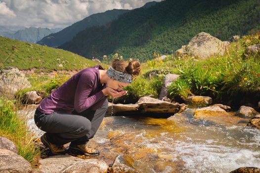 A woman drinks water from a mountain spring, sitting, a clear river in the mountains on a sunny day.