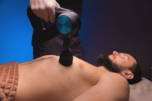 close-up of a professional male masseur massaging pectoral muscles with a percussion instrument, massage gun of a muscular athlete, in a spa treatment, lying on his back on a massage table.