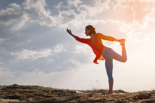 a woman practicing yoga in a pose stands on a rug on stones in the mountains against the background of clouds and the sun.