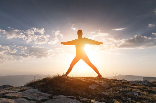 Silhouette of a happy woman at sunset or sunrise standing with her arms raised on top of a mountain while hiking or doing sports.