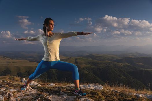 yoga warrior pose of young caucasian woman practicing at sunset in the mountains in the grass. Zen health and wellness concept.