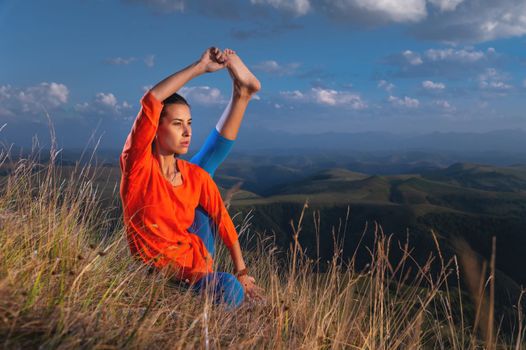 young woman practicing yoga poses raising one leg to her head while sitting on the rocks in the sunset light in the mountains.