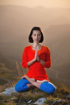 young caucasian woman meditates in the lotus position in the grass in the mountains near the cliff in the setting sun.
