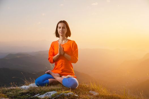 a yogi woman sits in a lotus position with crossed legs and holds her hands to her chest against the backdrop of a sunset in the mountains, there is a place for advertising on the side.