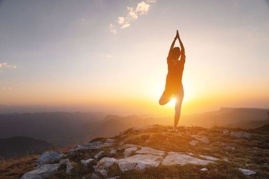 silhouette of a woman in a tree pose, yoga asana, stands near a cliff in the mountains against the backdrop of a sunset with a clear sky.