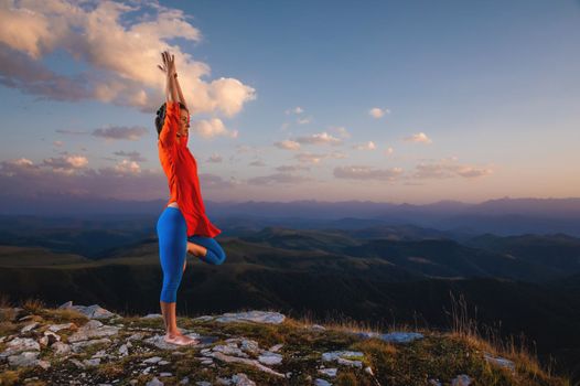 athletic woman stands in tree pose in yoga in the sunset light of the sun in the mountains.