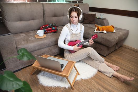 Beautiful young hipster girl in casual clothes learning to play the ukulele guitar while sitting on the floor in the living room at home, hobby leisure time concept.