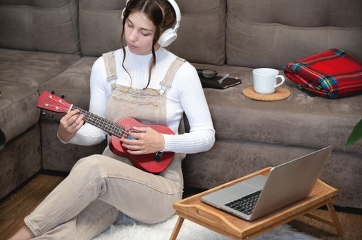 a young beautiful woman is sitting on the floor at home and learning to play the guitar or ukulele, standing next to a laptop and a cup of coffee. distance learning concept.