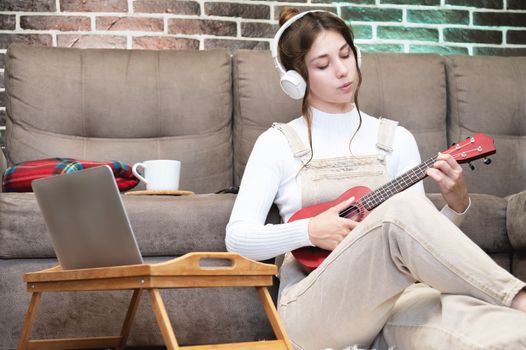 Beautiful young hipster girl in casual clothes learning to play the ukulele guitar while sitting on the floor in the living room at home, hobby leisure time concept.