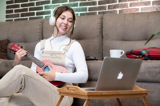 Beautiful young hipster girl in casual clothes learning to play the ukulele guitar while sitting on the floor in the living room at home, hobby leisure time concept. look at camera