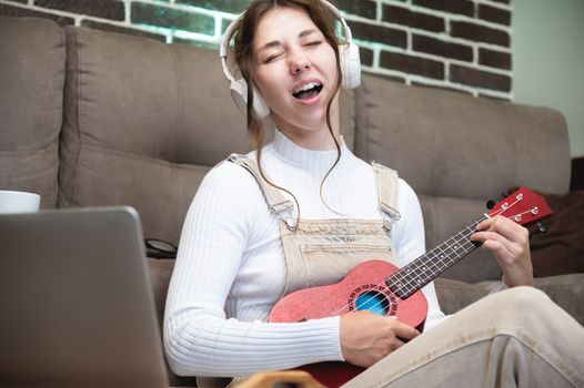 young woman student learning vocals and playing the ukulele at home sitting on the floor in the living room in front of a laptop.