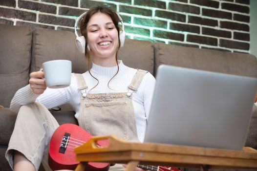 happy young woman sitting with a cup of coffee and ukulele, wearing headphones on the floor in the living room in her house, looking into the laptop monitor, distance learning.