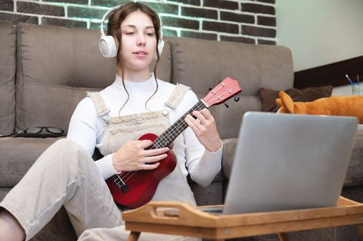 a young beautiful woman is sitting on the floor at home and learning to play the guitar or ukulele, standing next to a laptop and a cup of coffee. distance learning concept.
