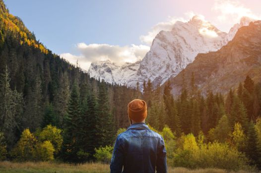 Back view of a man in a knitted hat and denim jacket against the backdrop of a green dense forest and snow-capped high mountains.