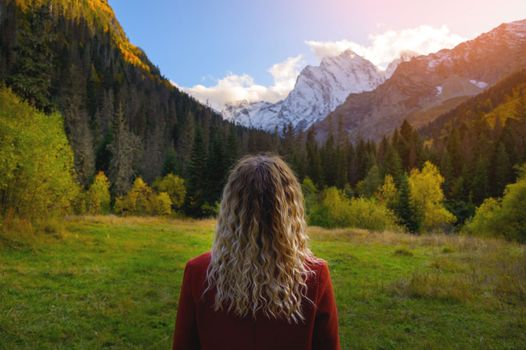 rear view, a blonde woman in a red coat stands against the backdrop of snow-capped mountains and a dense green forest.