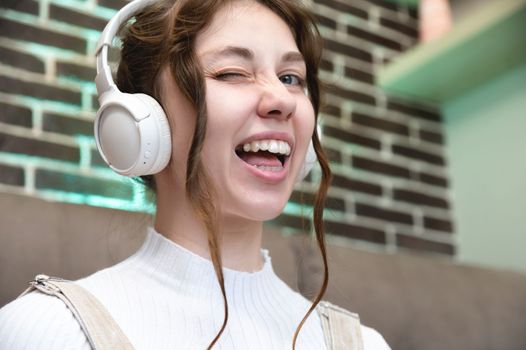 Young caucasian teenage woman smiles and winks, she listens to music in headphones while sitting at home.