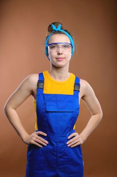 Woman builder in blue protective overalls and goggles standing and smiling, studio shot.
