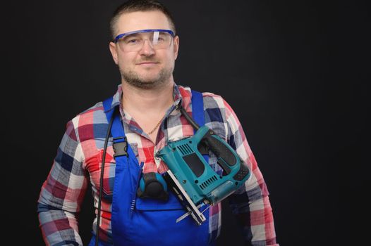 A male builder in goggles and a uniform on a black background smiles, a tool for work hangs around his neck. Studio shot, portrait.