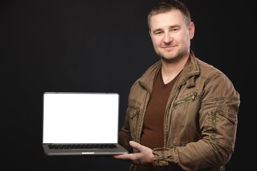 Smiling unshaven business man in a leather jacket on a black wall background. Achieving career wealth business concept. Holding a laptop computer with a blank screen.