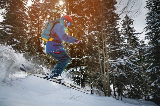 a skier makes a ski jump with a plume of snow behind him, in the mountains against the backdrop of a dense forest. male athlete in bright clothes makes a downhill race.