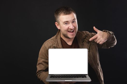 Portrait of a positive bearded man in casual clothes holding a laptop and pointing at a blank screen, excited, happy with a computer application. place for advertising.