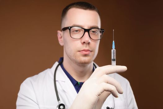 Caucasian male doctor holding a syringe in a bathrobe with a stethoscope and gloves on a plain background, pensive look, studio shot.