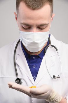 Portrait of a confident male doctor wearing a uniform with a stethoscope and a mask, holding pills in his palm and looking at them.