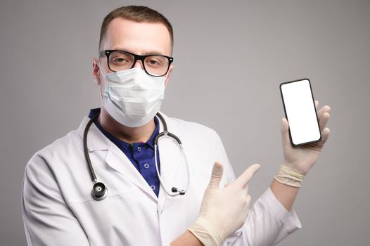 a young doctor in glasses and a mask stands pointing at the smartphone with his finger, looking at the camera, a portrait in a bathrobe.