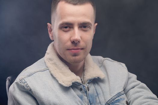 portrait of a young man in a denim jacket, looking straight at the camera, haze around, studio shot.