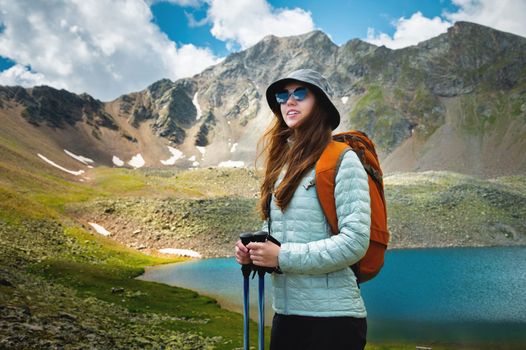 a young caucasian woman with a backpack and in a panama, stands with trekking poles near a mountain lake and looks into the distance. hiking, walking.