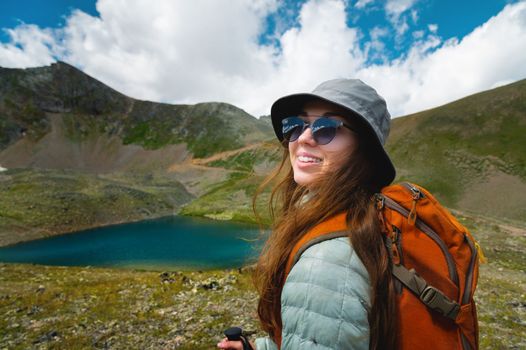 Beautiful young woman with a tourist backpack in a sports jacket and hat. Summer landscape of lake in green mountains.