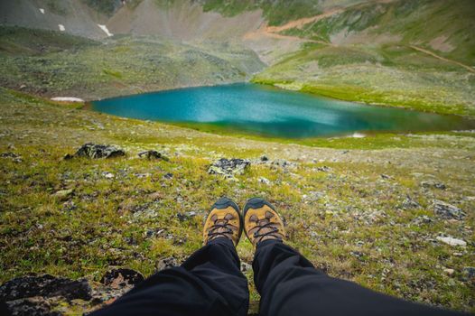 The traveler is resting on a mountain plateau. First-person view, close-up legs against the background of a mountain turquoise lake.