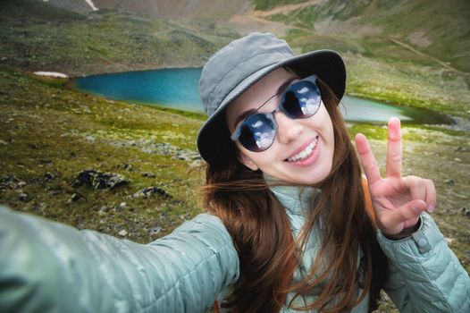 Young happy smiling tourist woman with long brunette hair and gray panama takes selfie with smartphone making peace sign with hand on top of rocky mountains with lake in background. Adventure, travel, vacation concept.