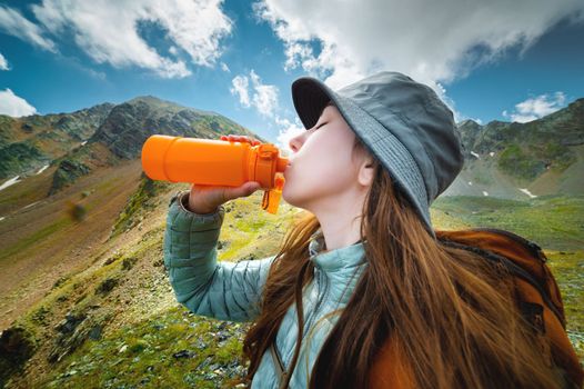 Young sports girl stands against the backdrop of a stunning mountain view and drinks clean water from a rubber bottle, trekking with a backpack.