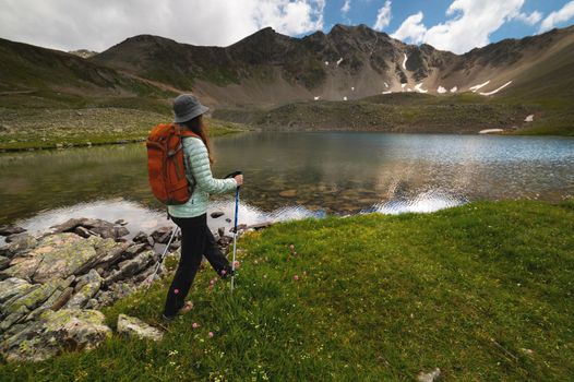 A woman with an orange backpack and a hat goes to a transparent lake in the mountains on a clear day.