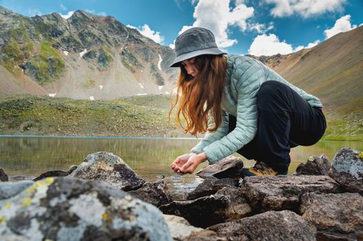 A young girl fills her hands with water from a stream of a cold mountain lake in order to drink and refresh herself. Happy hiker on a hike.