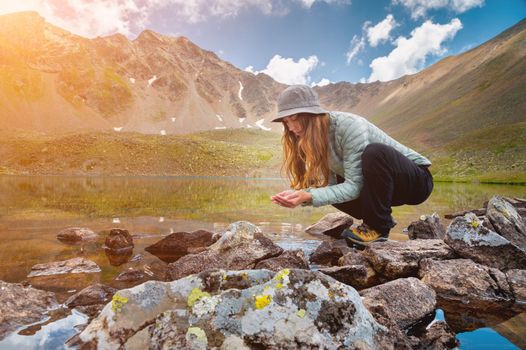 A young girl fills her hands with water from a stream of a cold mountain lake in order to drink and refresh herself. Happy hiker on a hike.